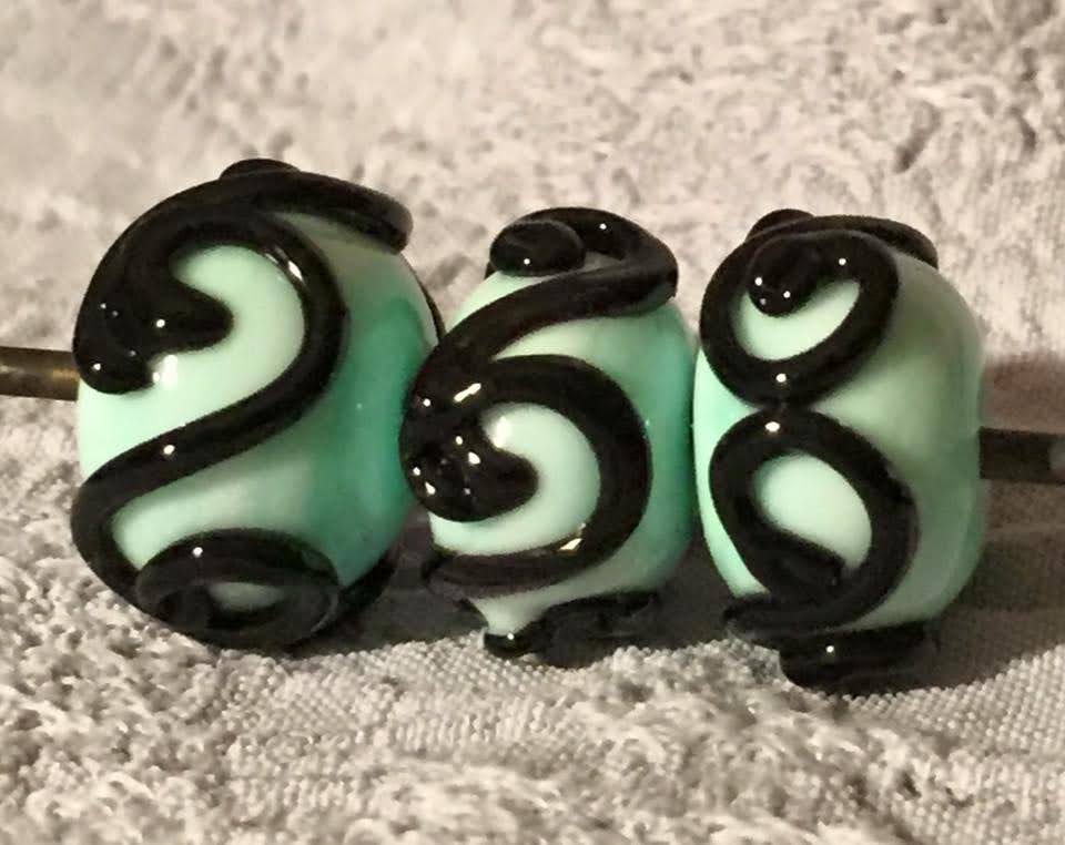 Three mint green glass beads showing black glass scrollwork.